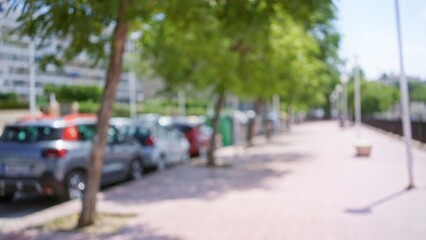 Outdoor scene in a sunny city with blurred cars parked along a street lined with trees and a sidewalk.