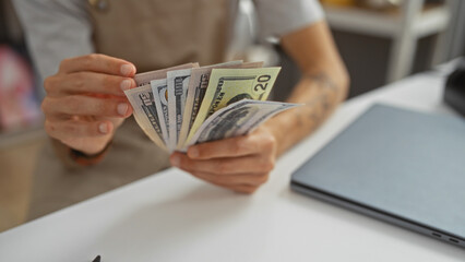 Young hispanic man counting us dollars in a store interior surrounded by home decor with a laptop on the desk