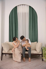 Young parents are playing with their 10 -month -old son on the sofa, Green curtain at the back