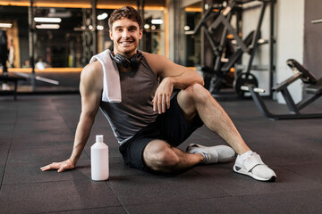 Sportive strong man taking break during workout day in gym, sitting with towel on shoulders and smiling at camera