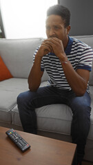 Young man sitting on couch in living room, hands clasped, thoughtful expression, remote on table, casual outfit, relaxed setting, natural light, indoors, home environment.