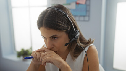 Woman working in office focused on task while wearing headset and holding pen indoor, workplace...