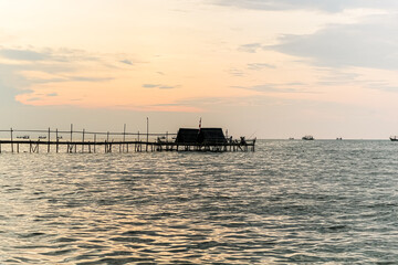 A dramatic sunset casts a warm glow on the calm waters, silhouetting a wooden structure extending into the sea. The sky is painted with hues of orange and pink.