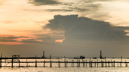 A dramatic sunset casts a warm glow on the calm waters, silhouetting a wooden structure extending into the sea. The sky is painted with hues of orange and pink.