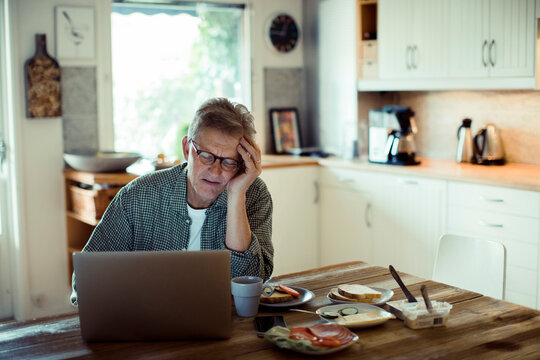 Stressed senior man using laptop on kitchen desk