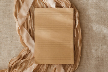 Blank lined sheet of paper card mockup, beige fabric and ribbon, top view on beige background with copy space. Flat lay, greeting card template, invitation. Neutral colours, aesthetic composition