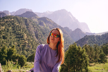 a young girl with loose red hair and glasses is sitting against the background of mountains. mountain trekking in summer