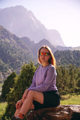 Naklejka premium a young girl with loose red hair and glasses is sitting against the background of mountains. mountain trekking in summer