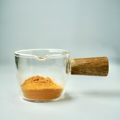 Turmeric powder in glass cup container with wooden handle isolated on square background.