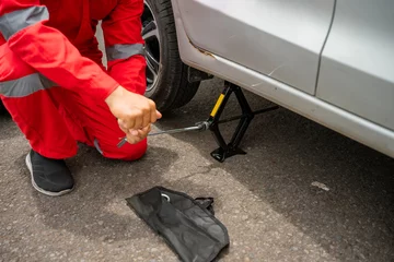 A mechanic using a jack to change a tire on a silver vehicle in an outdoor setting.