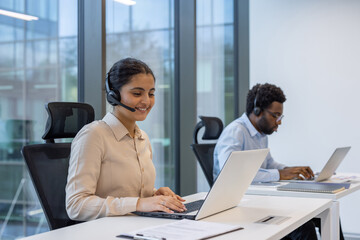 Two professionals in a modern office environment using laptops and headsets for communication. They...