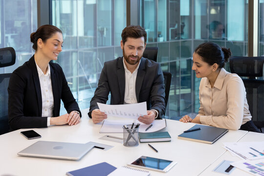 A professional office team is engaged in reviewing a financial report during a meeting. The group, dressed in business attire, collaborates over documents and utilizes digital devices