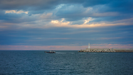 White lighthouse on the breakwater, rough waves, a  fisher boat and rough dark clouds