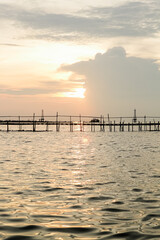 A dramatic sunset casts a warm glow on the calm waters, silhouetting a wooden structure extending into the sea. The sky is painted with hues of orange and pink.