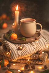 Steaming cup of hot beverage sits on a tray with a mandarin orange and lit candles, placed on a cozy knitted blanket with warm, inviting lighting, creating a perfect winter atmosphere