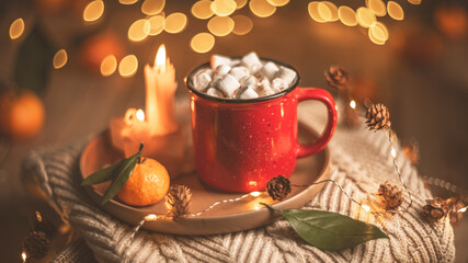 Red mug filled with hot chocolate and marshmallows resting on a tray with tangerines, surrounded by warm knit fabric and glowing christmas lights, creating a cozy winter scene