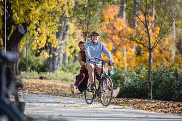 Smiling senior couple riding a bike in a sunny autumn park, wearing protective helmets. The man pedals while the woman enjoys the ride sitting on the rear rack