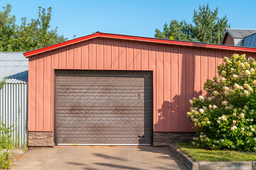 Fototapeta premium Bright and inviting garage with a red roof stands against a clear blue sky, bordered by lush green trees and flowering shrubs in full bloom