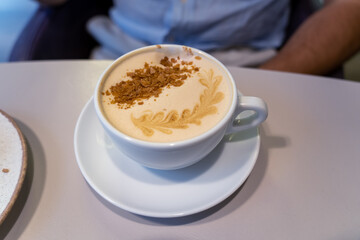 A creamy latte topped with decorative foam and crushed biscuits is presented on a saucer at a cozy cafe during midday hours