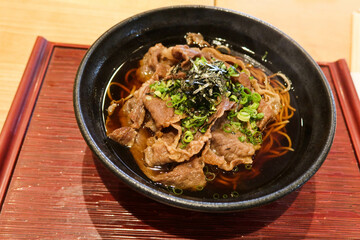 close up of hot Japanese beef soup soba noodles in a black bowl with wooden tray with spring onion on top