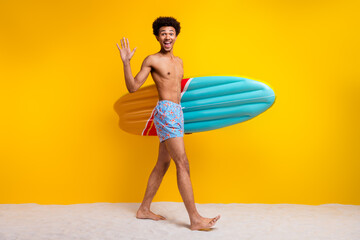 Happy young man with surfboard in summer beach attire posing against vibrant yellow background.