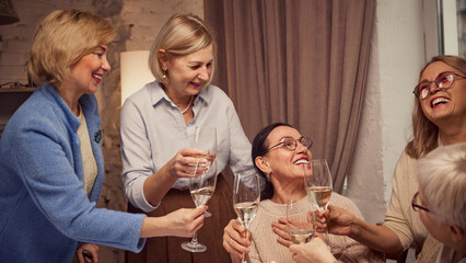 Birthday celebration. Group of elderly women, friends meeting at home in cozy living room, celebrating birthday of friend, drinking wine, saying toasts. Concept of elderly lifestyle, positive emotions