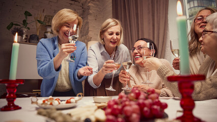 Lively cozy meeting. Elderly beautiful women meeting at home, cheerfully talking, laughing and drinking wine. Happiness and joy. Concept of elderly lifestyle, positive emotions