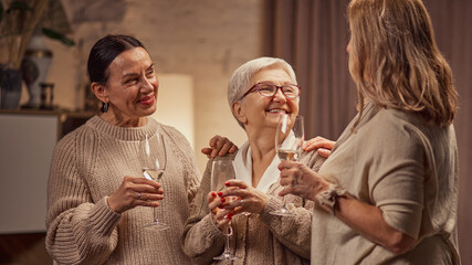 Happy, smiling elderly woman dressed in warm pastel clothes, meeting at home, in cozy living room, drinking wine, talking and laughing. Concept of elderly lifestyle, positive emotions