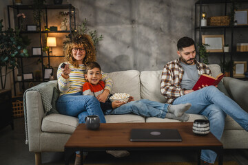Naklejka premium Mum, and son watch television and sit on sofa, while dad read a book