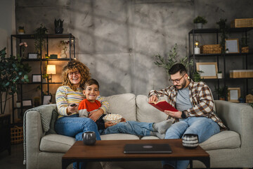 Naklejka premium Mum, and son watch television and sit on sofa, while dad read a book