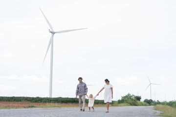 Obraz premium Father, mother and little daughter walking and holding hands together at wind turbine farm station. Happy smiling young mother with father and cute little daughter at windmill field farm