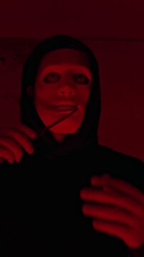Vertical lowkey medium portrait of mad Caucasian maniac taking off mask and licking sharp knife, passing it over face and neck, posing in abandoned room with flashing red light