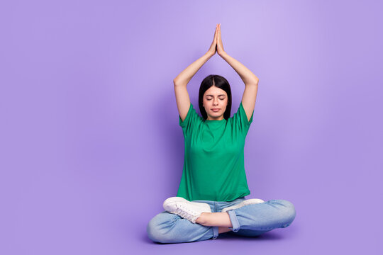 Young woman in casual outfit practices yoga with hands raised on a purple background