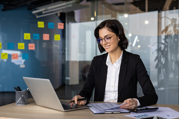 Confident businesswoman at desk using laptop and holding documents, engaged in work. Bright sticky...