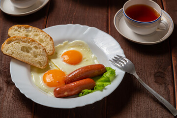 Breakfast fried eggs with sausages white bread and tea in a cup on a wooden table. Horizontal photo.