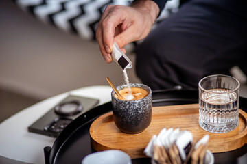  A hand adding sugar to a cup of espresso on a wooden tray with a glass of water. Highlights sugar's role in balancing coffee's flavor but also its health implications when consumed excessively.