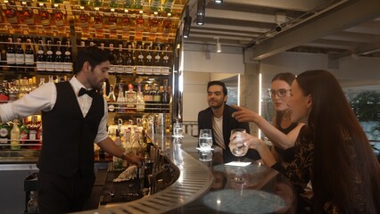 Smart bartender with VIP customer at luxurious bar greeting new beautiful guest at counter in engaging lively conversation at back view in lighting atmosphere at lighting warm nightclub. Vinosity.