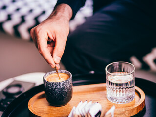 Description: Close-up of a person stirring a cup of espresso on a wooden tray with a glass of...