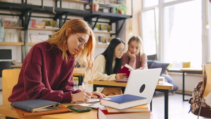 Red-haired female student in glasses sits in the foreground during an online lecture, taking notes in a notebook. Classmates are blurred in the background. - Powered by Adobe