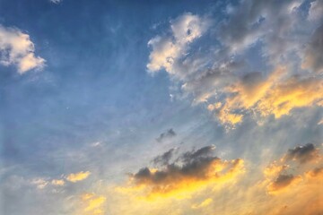 A bright sky at sunrise or sunset, with soft gradients of blue mixed with gold and orange, as clouds spread out, some illuminated by sunlight.