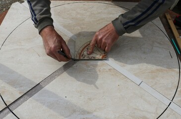 Hands of a craftsman with a pencil and a decorative tile fragment create a pattern of ceramic material on a round tabletop, tile decoration of individual furniture