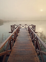 Obraz premium Wooden pier by the lake leading into a foggy scene, surrounded by tall dry reed. The sunrise sun is faintly visible through the mist, creating a serene and tranquil atmosphere. Calm and ethereal scene