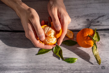 The beautiful hands of a young man are peeling a tangerine on a wooden table, next to the table there is a whole tangerine with a twig and leaves. Close-up.