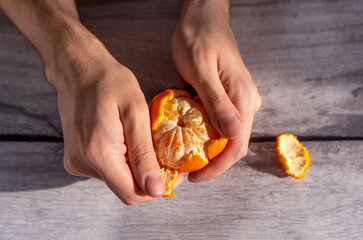 Beautiful hands of a young man peeling a tangerine on a gray wooden table.