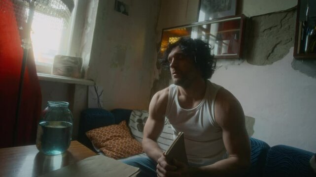 Full shot of apathetic and unhappy gen Y man in undershirt and jeans getting up and sitting on sofa, holding notebook and thinking about meaninglessness of life, living alone with little goldfish