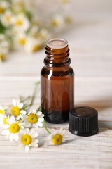Bottle of essential oil and chamomile flowers on white wooden table, closeup