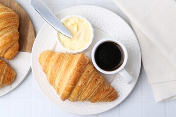 Tasty fresh croissants served with butter and cup of coffee on white tiled table, flat lay