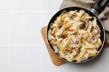 Tasty Alfredo pasta with chicken and mushrooms in frying pan on white tiled table, top view. Space for text