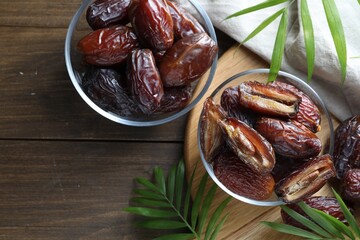 Many tasty dried dates in bowls and leaves on wooden table, flat lay