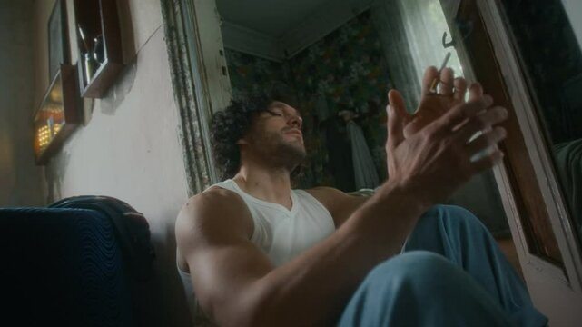 Medium shot of depressed and tired Caucasian man in undershirt and jeans sitting on floor in shabby house, smoking cigarette and thinking about hopeless future